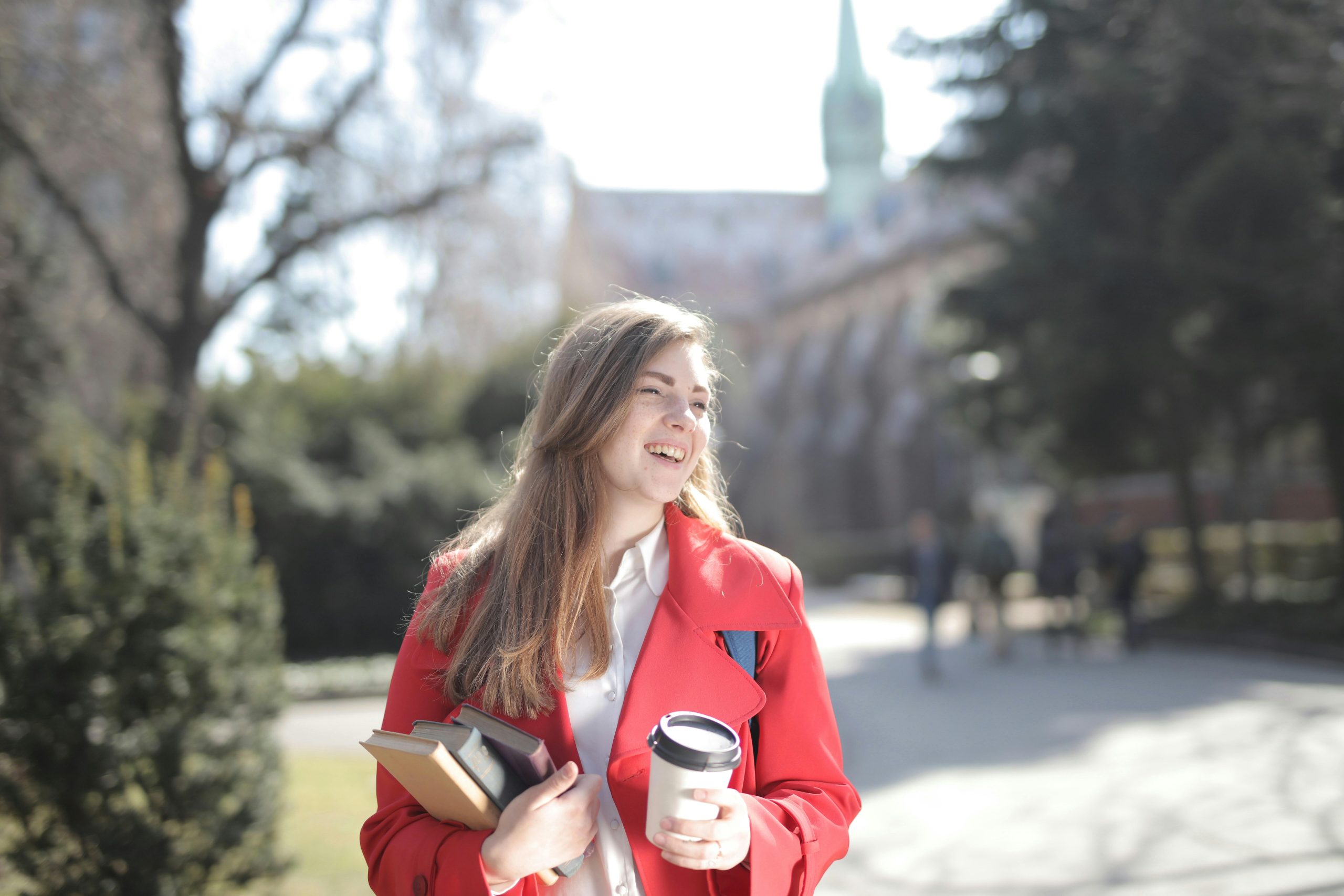 A cheerful young woman holding coffee and books, walking in a sunny campus setting.