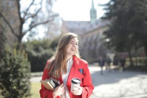 A cheerful young woman holding coffee and books, walking in a sunny campus setting.